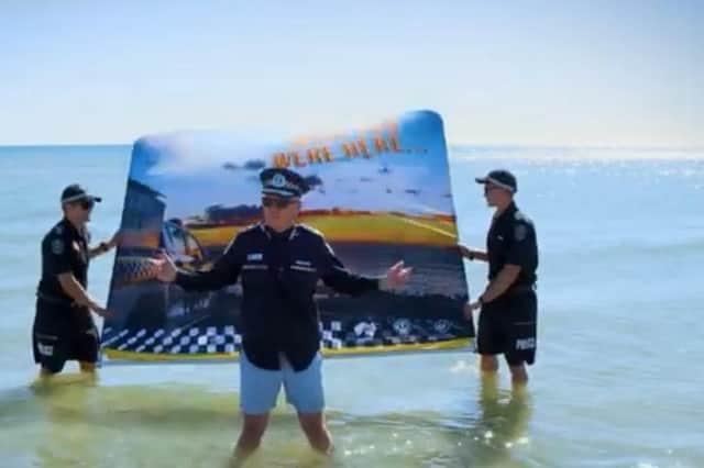 The video then zooms out to show the media wall fall backwards, revealing the Commissioner standing in the water at a picturesque South Australian beach, in his swimming shorts.